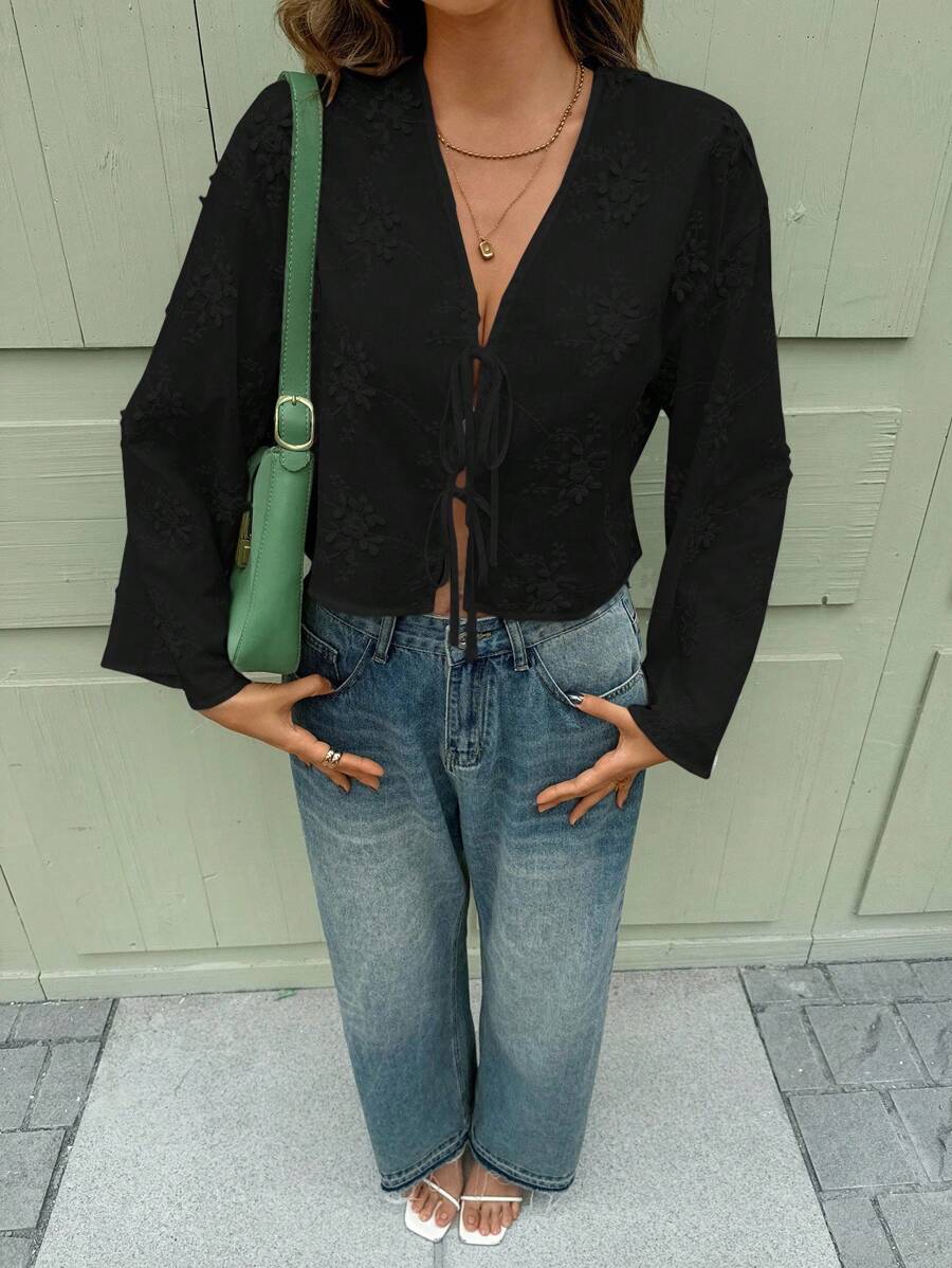 Person wearing a black top and blue jeans with a green bag, standing against a light gray wall.