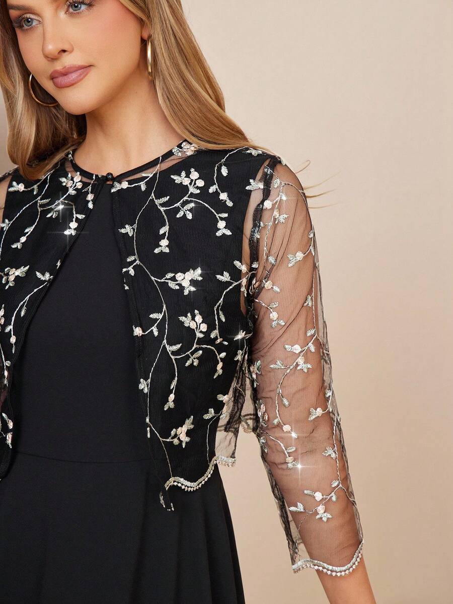 Woman wearing a black dress with sheer sleeves featuring floral embroidery on a beige background
